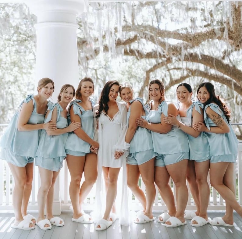 Peut inclure: Un groupe de femmes posent pour une photo, v&ecirc;tues de pyjamas bleu clair et de pantoufles blanches. La mari&eacute;e porte une robe et un peignoir blancs. La sc&egrave;ne se d&eacute;roule &agrave; l'ext&eacute;rieur, sur un porche blanc.