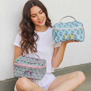 Pode incluir: Duas bolsas de cosméticos com estampas florais e alças. Uma é azul clara com flores roxas e iniciais bordadas em dourado. A outra é verde-azulado com flores rosas e iniciais bordadas em rosa. Ambas as bolsas têm formato retangular.