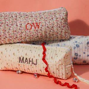 May include: Three quilted pencil cases with floral patterns. The top case is red and white, embroidered with "OW". The middle case is cream with "MAHJ". The bottom case is blue and white. Each has a tassel.