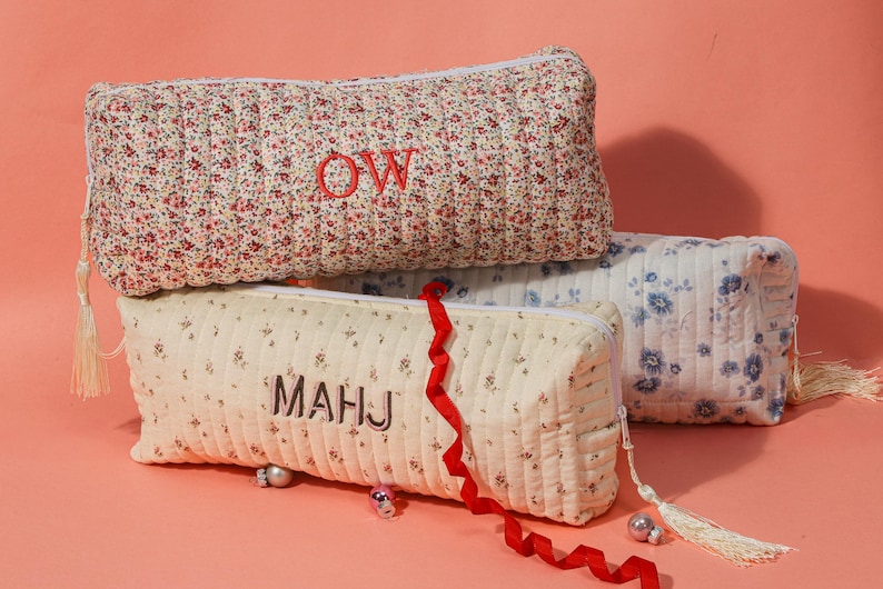 May include: Three quilted pencil cases with floral patterns. The top case is red and white, embroidered with "OW". The middle case is cream with "MAHJ". The bottom case is blue and white. Each has a tassel.