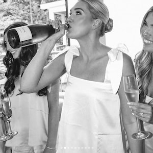 Peut inclure: Image en noir et blanc d'un groupe de femmes en f&ecirc;te. Une femme boit &agrave; une bouteille de champagne, tandis que d'autres tiennent des fl&ucirc;tes &agrave; champagne. Elles portent toutes des robes blanches. L'image est prise en ext&eacute;rieur.