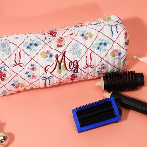 May include: A white quilted cosmetic bag with a floral and bow pattern, personalized with the name "Meg" in burgundy script. Two black hairbrushes with wooden handles and a blue-framed brush are next to the bag.