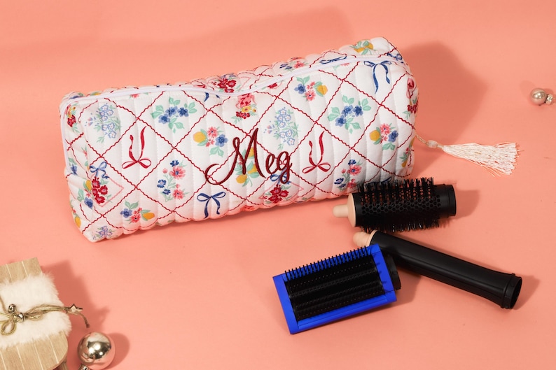 May include: A white quilted cosmetic bag with a floral and bow pattern, personalized with the name "Meg" in burgundy script. Two black hairbrushes with wooden handles and a blue-framed brush are next to the bag.