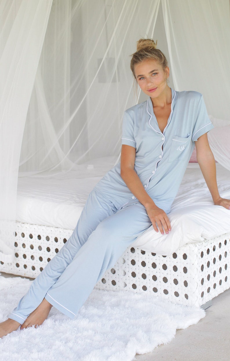 May include: A woman wearing a light blue pyjama set with white piping. The pyjamas have a button-up front and a pocket on the left side. The pyjamas are embroidered with the initials "A.S.B."