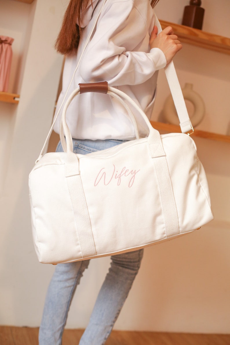 May include: White canvas duffel bag with a brown leather handle and a pink embroidered word "Wifey" on the front.