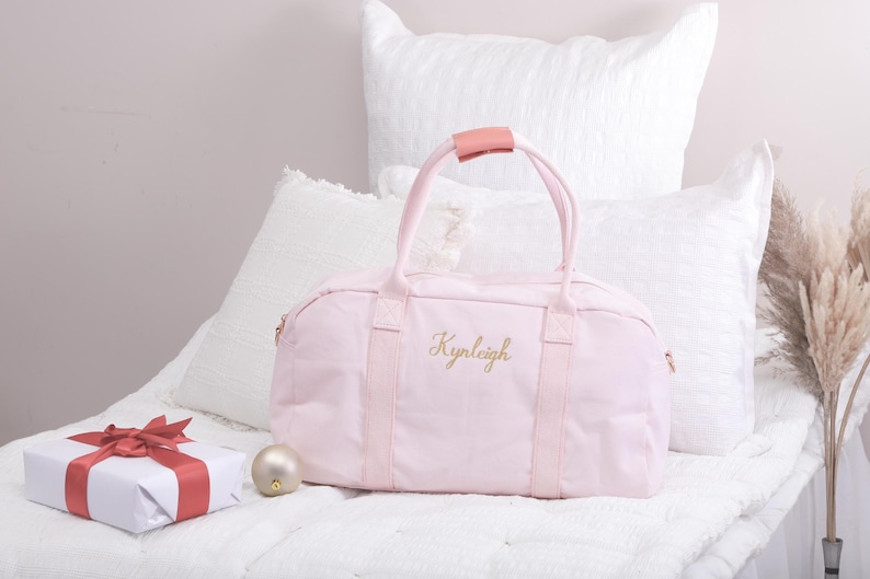 May include: A pink canvas duffel bag with a pink leather handle and the name "Kynleigh" embroidered in gold thread.