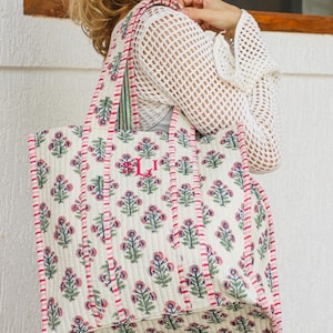 Puede incluir: Bolso tote acolchado grande con un estampado floral en rosa y verde sobre un fondo blanco roto. El bolso tiene ribetes y asas rosas, y un monograma. Dise&ntilde;ado para transportar objetos.