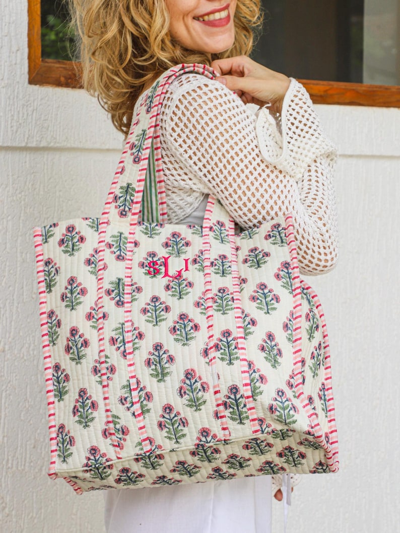 Puede incluir: Bolso tote acolchado grande con un estampado floral en rosa y verde sobre un fondo blanco roto. El bolso tiene ribetes y asas rosas, y un monograma. Dise&ntilde;ado para transportar objetos.
