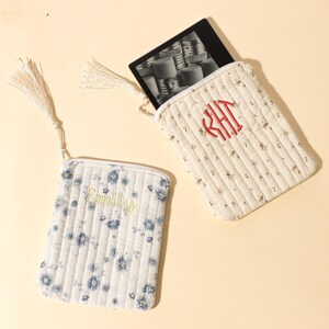 Puede incluir: Dos bolsas de tela acolchada con cremalleras y borlas. Una bolsa es blanca con estampados florales azules y el nombre "Emily" bordado en amarillo. La otra es de color crema con estampados florales rojos y bordado de monograma rojo.