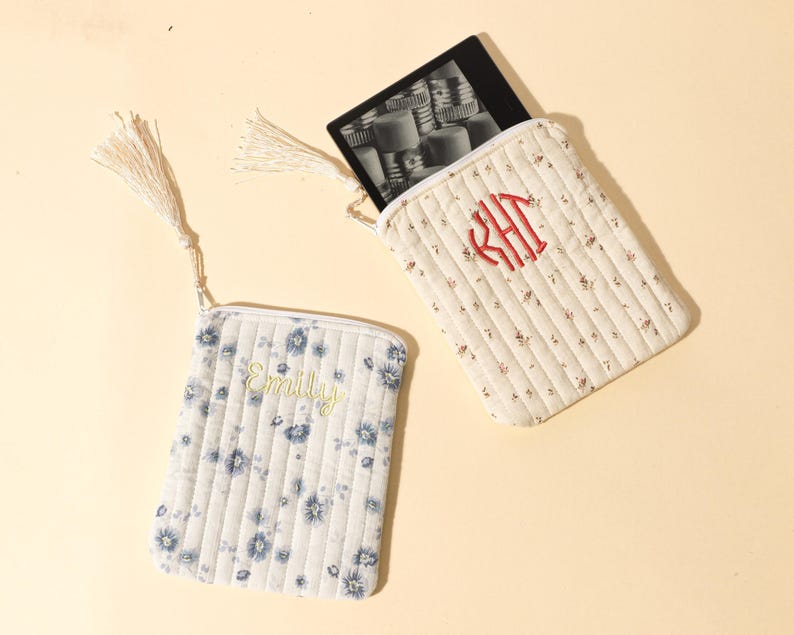 Puede incluir: Dos bolsas de tela acolchada con cremalleras y borlas. Una bolsa es blanca con estampados florales azules y el nombre "Emily" bordado en amarillo. La otra es de color crema con estampados florales rojos y bordado de monograma rojo.