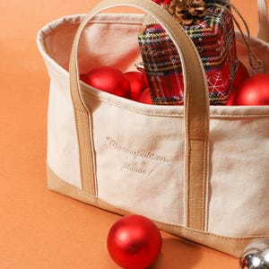 May include: A cream tote bag with tan handles and trim, embroidered with "Cosmopolitan please!". The bag contains red Christmas baubles and a wrapped gift with a plaid pattern. Some baubles are scattered around the bag.