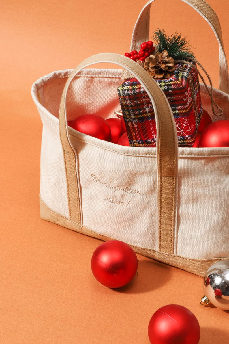 May include: A cream tote bag with tan handles and trim, embroidered with "Cosmopolitan please!". The bag contains red Christmas baubles and a wrapped gift with a plaid pattern. Some baubles are scattered around the bag.