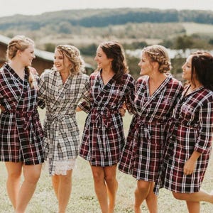 May include: Five women wearing plaid robes with embroidered initials. The robes are in shades of red, black, and white. The women are standing in a field with green grass and a blurry background.