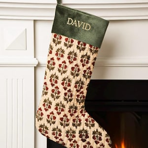 May include: A Christmas stocking with a green velvet cuff embroidered with the name "DAVID" in gold. The body of the stocking is cream-colored with a repeating pattern of red and green floral designs. A green loop is attached for hanging.