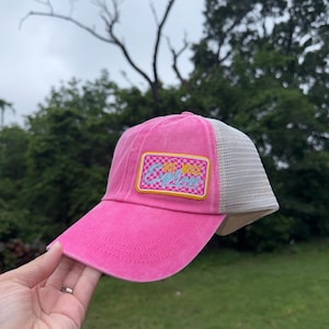 May include: A pink trucker hat with a mesh back and a curved brim. The hat features a rectangular patch with the words "BUT MESS" in a retro font, set against a checkered background. The hat is held by a person.