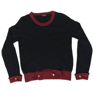 May include: A long-sleeved navy blue sweater with a red trim around the neckline and cuffs. The sweater has gold buttons on the cuffs and bottom hem.