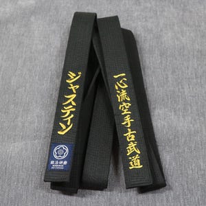 May include: A black martial arts belt featuring gold embroidered Japanese characters on both ends. One end displays a small blue rectangular patch with a white floral emblem and the text BERSERKER Belt Craftsman in white lettering.
