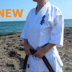 Könnte beinhalten: Weißer Karateanzug mit schwarzem Gürtel. Der Anzug hat ein traditionelles Design mit einem V-Ausschnitt-Oberteil und einer Hose mit weitem Bein. Der Text "NEW" wird in Orange angezeigt. Der Hintergrund ist eine felsige Küstenlinie mit dem Meer.