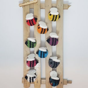 Op de afbeelding: Een houten lattenrek met negen gehaakte hommels in verschillende kleuren, waaronder rood, oranje, geel, groen, blauw, paars, roze, grijs en teal. Elke hommel heeft een witte gehaakte vleugel.