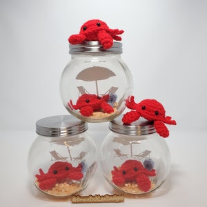 May include: Three glass jars with silver lids, each containing a red crocheted crab and a miniature beach scene. The top jar has a crab on top of the lid. The jars are stacked on top of each other. The bottom jar has the text "@GraceDuhLeeDesigns" on the front.