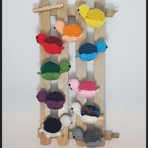 Peut inclure: Collection de neuf figurines de tortues crochetées colorées fixées sur une clôture en bois. Les tortues sont de différentes couleurs, dont le rouge, le jaune, le vert et le bleu. La clôture est de couleur marron clair et est montée sur un fond blanc. Le nom de l'artiste, "GraceDulLeeDesigns", est visible.