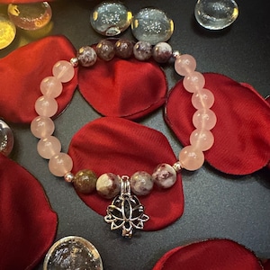 Könnte beinhalten: Ein rosa-braunes Perlenarmband mit einem silbernen Anhänger in Form einer Lotusblume. Das Armband liegt auf einem schwarzen Hintergrund mit roten Rosenblättern.