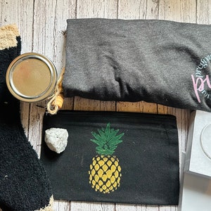 Könnte beinhalten: Ein Geschenkkorb mit verschiedenen Artikeln, darunter ein graues T-Shirt mit dem Text "purpo" in Pink, eine schwarz-weiße Socke, eine schwarze Kosmetiktasche mit Ananas-Motiv, ein weißer Stein, ein Glasgefäß und eine weiße Schachtel mit zwei silbernen Creolen.