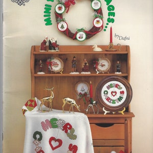 May include: A book titled "Mini Yuletide Messages" by Dafni, featuring a collection of cross-stitch patterns for Christmas ornaments and decorations. The cover shows a wreath with ornaments, a shelf with figurines, and a table with a cross-stitch design of a heart with the words "Merry Christmas".