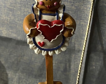 May include: A gingerbread chef ornament holding a red heart. The ornament features a white chef's hat and apron with blue trim. The ornament is attached to a gold-colored string for hanging.