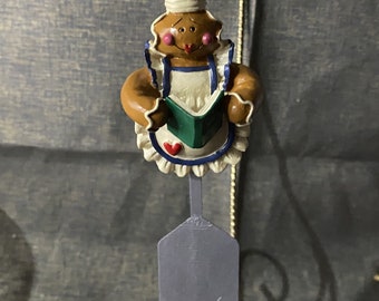 May include: Gingerbread person Christmas ornament with a white chef hat, holding a green cookbook and wearing a blue and white apron. The ornament is hanging from a gold string and has a gray spatula with red hearts on the bottom.