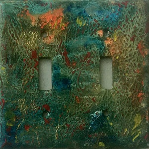 May include: A decorative double light switch cover with an abstract design. The cover has a textured surface with teal, orange, red, and yellow. The two switch openings are rectangular.