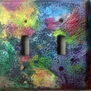 May include: A double light switch cover with a colourful abstract design. The cover has a textured surface with a vibrant mix of colours, including yellow, blue, red, and green. Two light switches are visible in the centre.