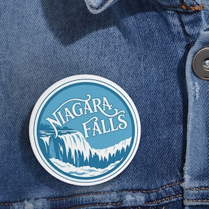 May include: A circular pinback button featuring a blue and white graphic of Niagara Falls with the text Niagara Falls in a stylized white font. The button is attached to a blue denim jacket with visible metal buttons and stitching details.