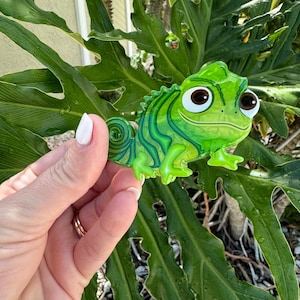 May include: A green chameleon figurine with a cartoonish design. The figurine has large, expressive eyes and a curled tail. The chameleon is held in front of a backdrop of large green leaves.
