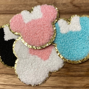 May include: Three felt patches in the shape of a cartoon character with a bow. The patches are pink, blue, and white with gold glitter trim.