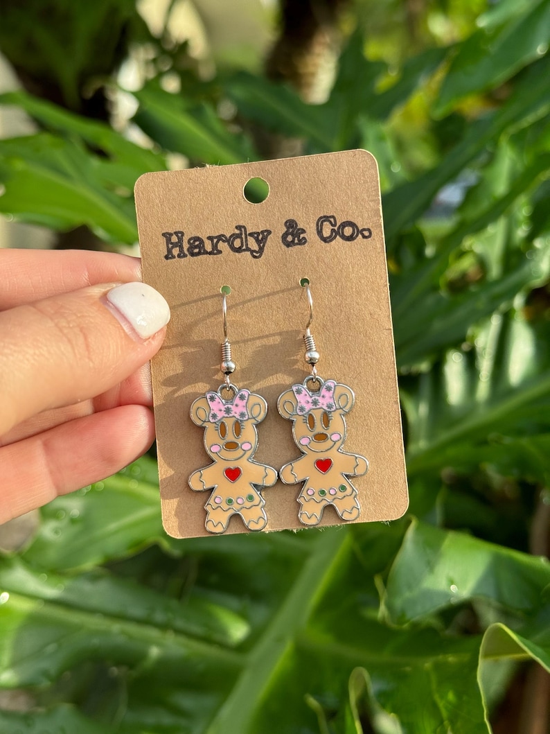 May include: A pair of silver dangle earrings featuring gingerbread person charms with pink bows, red hearts, and green accents. The earrings are displayed on a brown card with the text "Hardy & Co."