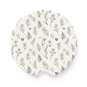 May include: A round, off-white car coaster featuring a repeating botanical pattern of black line-drawn flowers, leaves, and ferns, interspersed with small, subtle black silhouettes of three-circle mouse head shapes throughout the design.