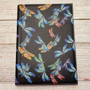 Puede incluir: Una libreta de cuero negro con un patrón de libélulas coloridas. Las libélulas son de color azul, verde y naranja iridiscente.