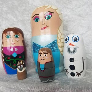 May include: A set of five hand-painted wooden nesting dolls featuring characters from the movie Frozen. The largest doll is a blonde woman with a blue dress and a white cape. The second largest doll is a brunette woman with a pink and blue dress. The third largest doll is a boy with a blue shirt and gray pants. The fourth largest doll is a snowman with a black top hat and a carrot nose. The smallest doll is a brown and black creature with a white face.