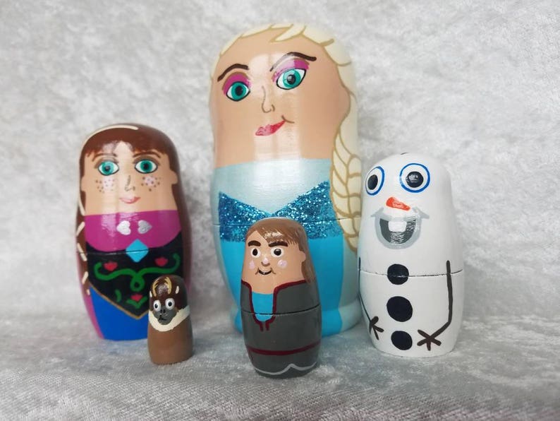 May include: A set of five hand-painted wooden nesting dolls featuring characters from the movie Frozen. The largest doll is a blonde woman with a blue dress and a white cape. The second largest doll is a brunette woman with a pink and blue dress. The third largest doll is a boy with a blue shirt and gray pants. The fourth largest doll is a snowman with a black top hat and a carrot nose. The smallest doll is a brown and black creature with a white face.
