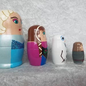 May include: Set of five hand-painted wooden nesting dolls featuring characters from the animated film Frozen. The largest doll is a light blue dress with a white bodice and blonde hair. The second largest doll is wearing a purple and green dress with brown hair. The third largest doll is white with a black face and a black arm. The fourth largest doll is wearing a gray and brown outfit with brown hair. The smallest doll is brown with a brown hat.