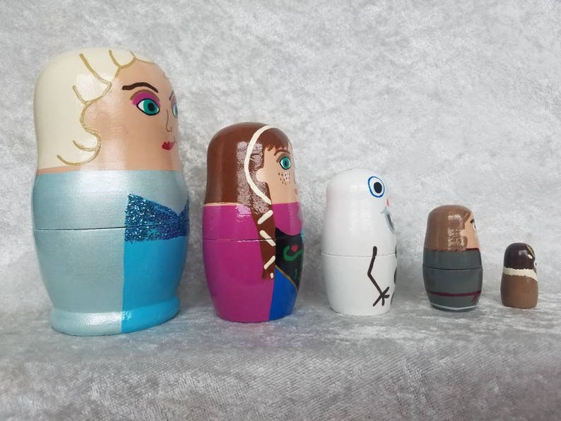 May include: Set of five hand-painted wooden nesting dolls featuring characters from the animated film Frozen. The largest doll is a light blue dress with a white bodice and blonde hair. The second largest doll is wearing a purple and green dress with brown hair. The third largest doll is white with a black face and a black arm. The fourth largest doll is wearing a gray and brown outfit with brown hair. The smallest doll is brown with a brown hat.