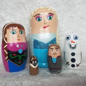 May include: Set of five hand-painted wooden nesting dolls featuring characters from the animated film Frozen. The dolls are painted in bright colors and depict Elsa, Anna, Olaf, Sven, and Kristoff.