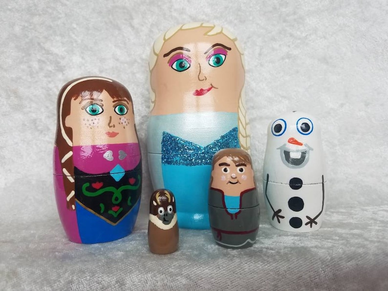 May include: Set of five hand-painted wooden nesting dolls featuring characters from the animated film Frozen. The dolls are painted in bright colors and depict Elsa, Anna, Olaf, Sven, and Kristoff.