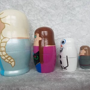 May include: Set of five hand-painted wooden nesting dolls featuring characters from the movie Frozen. The dolls are painted in various colors, including blue, white, pink, brown, and gray. The largest doll depicts Elsa with long blonde hair, the second largest doll depicts Anna with long brown hair, the third largest doll depicts Olaf the snowman, the fourth largest doll depicts Kristoff with brown hair, and the smallest doll depicts Sven the reindeer.
