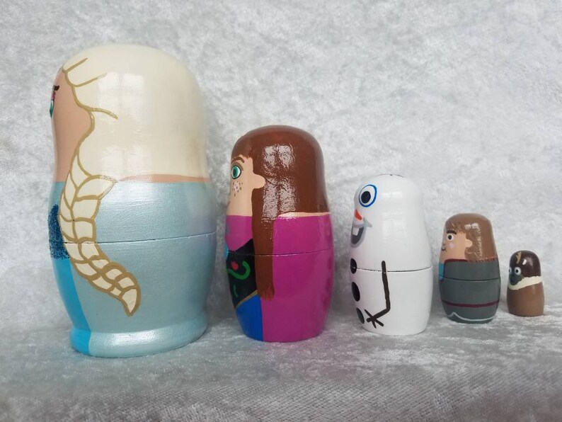 May include: Set of five hand-painted wooden nesting dolls featuring characters from the movie Frozen. The dolls are painted in various colors, including blue, white, pink, brown, and gray. The largest doll depicts Elsa with long blonde hair, the second largest doll depicts Anna with long brown hair, the third largest doll depicts Olaf the snowman, the fourth largest doll depicts Kristoff with brown hair, and the smallest doll depicts Sven the reindeer.