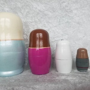 May include: Set of five nesting dolls, also known as Russian nesting dolls. The largest doll is light blue with a white top, the second largest is pink with a brown top, the third largest is white, the fourth largest is brown with a gray band, and the smallest is brown.
