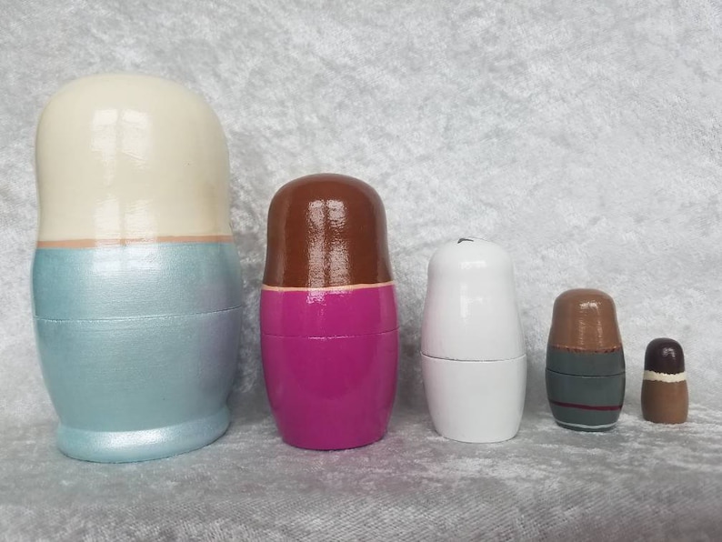 May include: Set of five nesting dolls, also known as Russian nesting dolls. The largest doll is light blue with a white top, the second largest is pink with a brown top, the third largest is white, the fourth largest is brown with a gray band, and the smallest is brown.