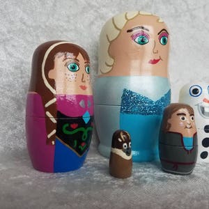 May include: Set of five hand-painted wooden nesting dolls featuring characters from the animated film Frozen. The dolls are painted in shades of blue, purple, brown, and white. The largest doll depicts Elsa, the second largest doll depicts Anna, the third largest doll depicts Olaf, the fourth largest doll depicts Kristoff, and the smallest doll depicts Sven.