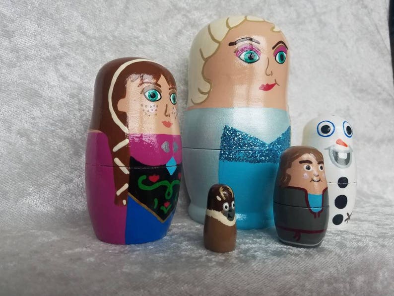 May include: Set of five hand-painted wooden nesting dolls featuring characters from the animated film Frozen. The dolls are painted in shades of blue, purple, brown, and white. The largest doll depicts Elsa, the second largest doll depicts Anna, the third largest doll depicts Olaf, the fourth largest doll depicts Kristoff, and the smallest doll depicts Sven.